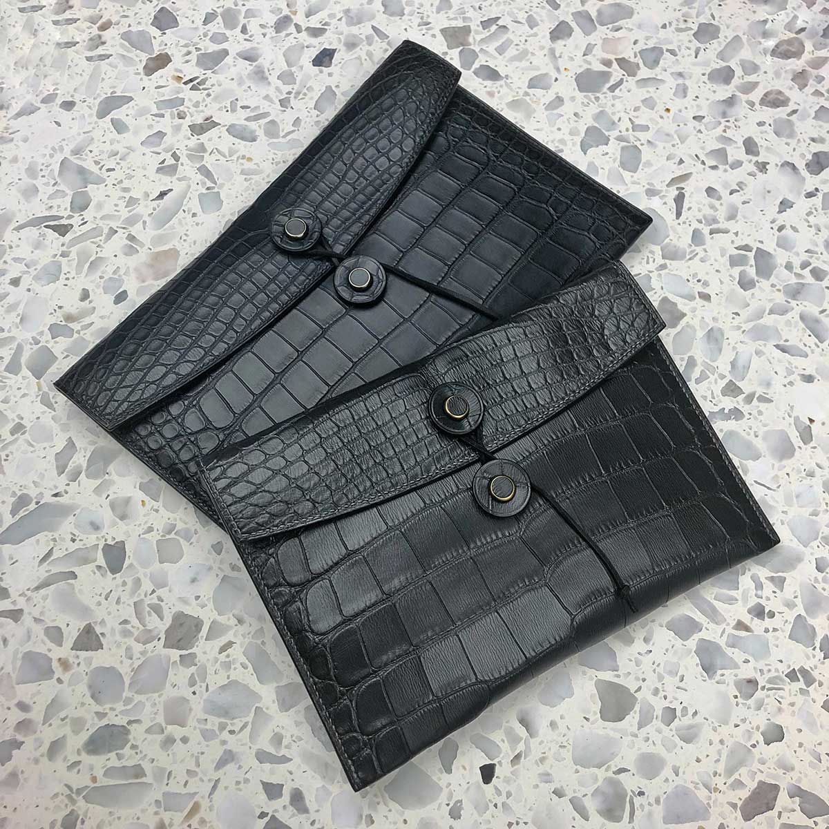 A black leather pouch with a crocodile pattern and multiple button closures, placed on a speckled surface.