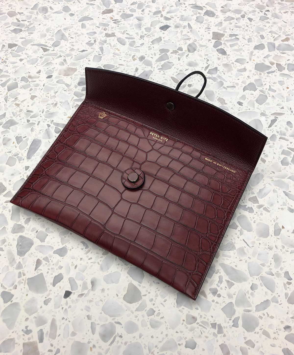 An open tether pouch in bordeaux alligator material laying on a terrazzo surface